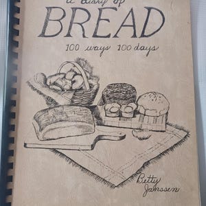 May include: A spiral-bound cookbook titled A diary of BREAD 100 ways 100 days by Betty Janssen. The tan cover features a black ink illustration of a basket of rolls, a loaf of bread on a cutting board, and various baked goods on a cloth.
