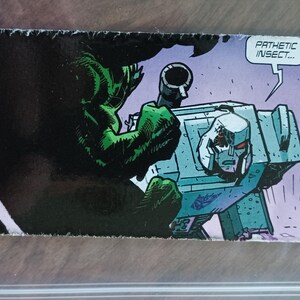 Pode incluir: Painel de banda desenhada com uma figura verde e musculosa dominando um personagem robótico azul claro com um rosto prateado. Um balão de fala contém o texto: "PATHETIC INSECT..."