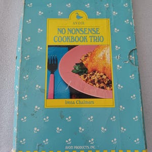 Puede incluir: Una caja vintage de Avon "No Nonsense Cookbook Trio". La caja es de color turquesa con un estampado floral y una etiqueta rectangular amarilla. La etiqueta presenta una imagen de una comida y el nombre "Irena Chalmers".