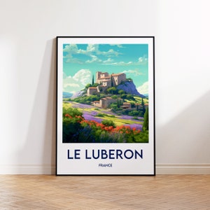 Könnte beinhalten: Gerahmter Kunstdruck mit einer farbenfrohen Landschaft von Le Luberon, Frankreich. Das Kunstwerk zeigt ein Dorf auf einem Hügel, umgeben von Lavendelfeldern und roten Blumen unter blauem Himmel mit Wolken. Der Druck hat einen schwarzen Rahmen.