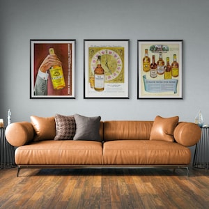 Puede incluir: Tres anuncios de whisky vintage enmarcados cuelgan sobre un sofá de cuero marrón. Los carteles presentan botellas de whisky y texto. El sofá tiene varios cojines y está sobre un suelo de madera.