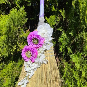 May include: A decorative broom with a natural straw head and a handle wrapped in white and lavender ribbon. The broom is adorned with purple and white flowers, and a sparkling rhinestone embellishment.