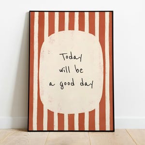 May include: A rectangular wall art print featuring a vertical striped pattern in terracotta and off-white. Centered is a large, rounded off-white shape containing the black handwritten text: Today will be a good day. The print is in a thin black frame.