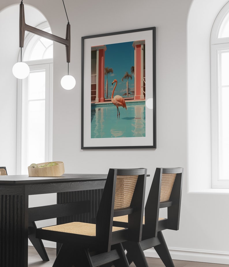May include: A black framed print of a pink flamingo standing in a blue pool with a pink building in the background.