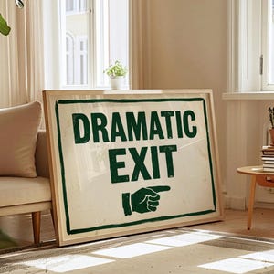 May include: A framed sign with the words "DRAMATIC EXIT" in dark green, with a pointing hand graphic. The sign has a light-coloured background and a wooden frame. The sign is leaning against a wall.