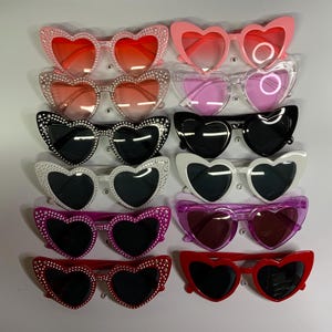 May include: A set of ten heart-shaped sunglasses with rhinestone accents. The sunglasses come in a variety of colors, including pink, red, black, white, and purple.