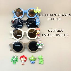 Op de afbeelding: Vier zonnebrillen in blauw, zwart, wit en grijs, elk met ronde monturen en donkere glazen. De zonnebrillen zijn versierd met diverse cartoon-personage versieringen. De tekst op de afbeelding luidt "30 VERSCHILLENDE GLASKLEUREN" en "MEER DAN 300 VERSIERINGEN."
