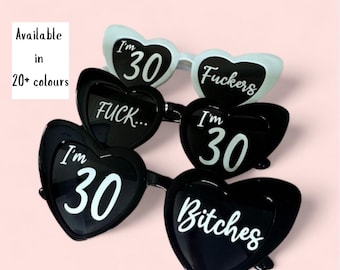 Könnte beinhalten: Herzförmige Sonnenbrillen in Schwarz und Weiß. Die Sonnenbrillen tragen die Aufschriften "I'm 30", "Fuckers", "Fuck..." und "Bitches". Das Bild enthält auch den Text "Available in 20+ colours".