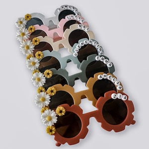 May include: A stack of colourful sunglasses with daisy embellishments and personalised names, including Samantha, Nayla, Mai, Clementine, Emma, Amelia, and Zia.