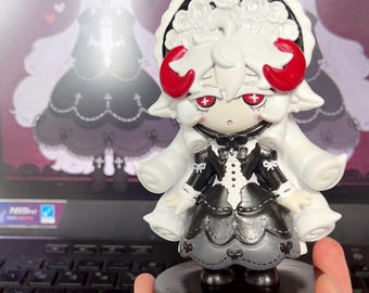 Könnte beinhalten: Eine kleine Figur, die einen Charakter mit weißem Haar, roten Hörnern und roten Augen darstellt. Der Charakter trägt ein schwarz-weißes Kleid mit einem rosenverzierten Kopfschmuck und steht auf einem runden schwarzen Sockel. Die Figur ist blass.