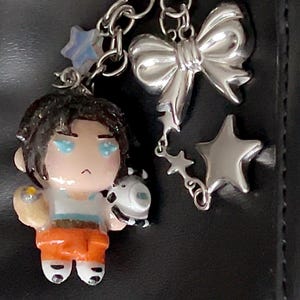 May include: A keychain featuring a small, hand-painted figurine with dark hair, a white top, and orange pants, holding a white and black spherical object. The charm is attached to a silver-toned chain with a large bow charm and two star charms.