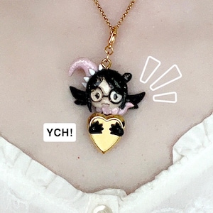 Puede incluir: Un collar con colgante dorado con un personaje de dibujos animados con cabello negro, gafas y alas negras, sosteniendo un corazón dorado. El personaje tiene detalles en rosa y está suspendido de una delicada cadena dorada. El texto "YCH!" está en un cuadro blanco.