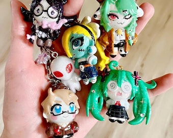 Puede incluir: Una colección de cinco pequeños dijes de personajes pintados a mano y con purpurina, unidos a un llavero de metal. Las figuras incluyen un personaje de pelo oscuro con gafas, una figura con cuernos y pelo verde, una figura de pelo amarillo, un fantasma blanco y una figura de pelo verde con coletas.