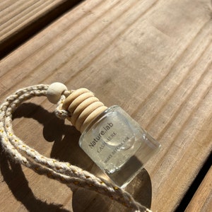 May include: A clear, square-shaped car air freshener with a wooden cap and a beige rope. The label reads "Nature.lab CASHMERE" with the instruction "SHAKE BEFORE USE". The rope has gold accents. The freshener sits on a wooden surface.