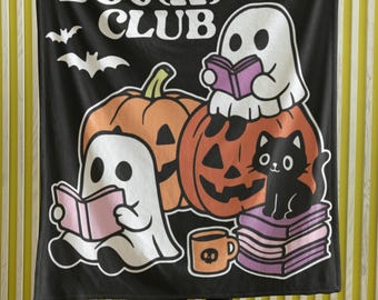 May include: Black blanket with a Halloween-themed design. Features the text "Midnight BOOK CLUB" with illustrations of ghosts reading books, pumpkins, a black cat, and a coffee mug. The blanket is held up by a person.