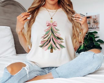 May include: Cream-colored t-shirt featuring a watercolor Christmas tree design. The tree has pink, green, and gold brushstrokes, topped with a pink bow and a gold star. The shirt is a casual, festive apparel item.