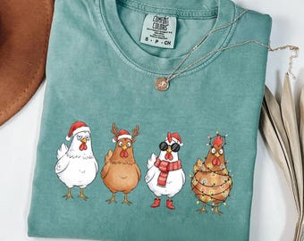 May include: A teal Comfort Colors t-shirt featuring four cartoon chickens in festive holiday attire. One chicken wears a Santa hat, another a reindeer antler headband, a third sports sunglasses and a scarf, and the last is wrapped in Christmas lights.