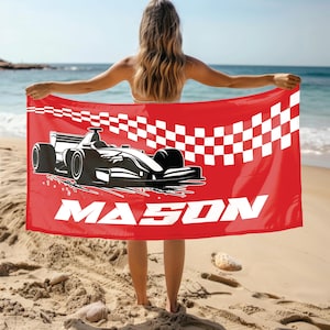 May include: Red beach towel featuring a black and white race car design and the name "MASON" in white block letters. The towel has a checkered flag pattern on the upper right corner. The towel is held up on a sandy beach.