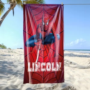 May include: A red beach towel featuring Spider-Man in a crouched pose against a spiderweb background. The towel has the name "LINCOLN" printed in white block letters. The towel is hanging on a clothesline at the beach.