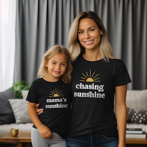 May include: Two people wearing black t-shirts with a yellow sun graphic and text. The adult shirt says "chasing sunshine" and the child's shirt says "mama's sunshine".