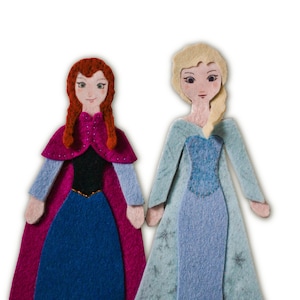 Puede incluir: Dos muñecas de fieltro, una con trenzas rojas, capa rosa y vestido azul, y la otra con cabello rubio, vestido celeste con detalles de copos de nieve. Ambas muñecas se toman de la mano.