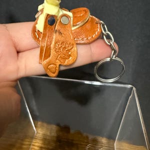 May include: A miniature brown leather saddle keychain with a silver keyring. The saddle has a cream-colored seat and detailed stitching. The keychain is a western-themed accessory.