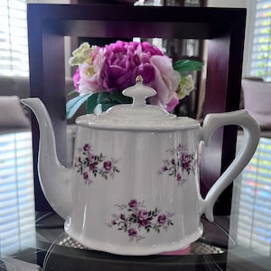 May include: White teapot with a floral design featuring purple roses and green leaves. The teapot has a gold lid knob and a curved handle.