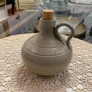May include: A gray ceramic bottle with a cork stopper. The bottle has a textured surface and a rounded body. It is sitting on a crocheted doily.