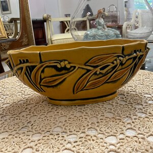 May include: A large, oval-shaped ceramic planter with a brown glaze and a design of stylized leaves and berries. The planter is sitting on a crocheted doily.