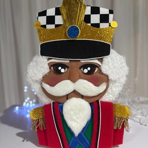 May include: A decorative nutcracker bust featuring a gold glitter crown with a black and white checkered band, a black brim, and a blue circular accent. The figure has white textured hair, a white mustache and beard, and a red, green, and blue uniform.