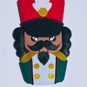 Puede incluir: Un adorno de soldado cascanueces de fieltro con un sombrero rojo, pelo negro y bigote negro. El soldado tiene una chaqueta verde con charreteras doradas y botones blancos. El diseño es brillante y festivo.