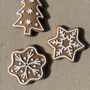 Puede incluir: Tres galletas de jengibre con forma de árbol de Navidad, copo de nieve y flor. Cada galleta es marrón con detalles de glaseado blanco. El árbol tiene decoraciones de estrellas y puntos.