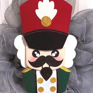 Puede incluir: Una decoración de cascanueces con un sombrero rojo, un penacho blanco y un detalle de purpurina dorada, un bigote negro y una chaqueta verde con cuatro botones de purpurina dorada. La figura tiene pelo blanco sobre un fondo de malla gris.