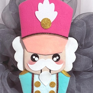 Puede incluir: Una decoración de cascanueces con un sombrero rosa brillante con un penacho blanco y un detalle de purpurina dorada, una chaqueta azul claro con botones de purpurina dorada, pelo blanco, bigote blanco, ojos negros grandes y mejillas rosadas.