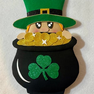 Pode incluir: Decoração do Dia de São Patrício com um duende espreitando por cima de um pote preto cheio de moedas de ouro. O duende usa um chapéu verde com uma faixa preta e uma fivela dourada. Um trevo verde brilhante está no pote.