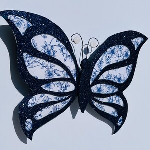 May include: A black glitter butterfly with white wings featuring a blue floral pattern. The butterfly has two white pearl accents on the antennae.