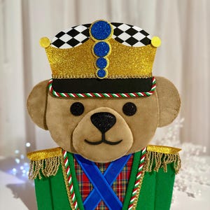 Può includere: Un orsacchiotto di peluche vestito da soldatino schiaccianoci, con una corona glitterata dorata a scacchi bianchi e neri, una giacca in feltro verde con spalline a frange dorate, un gilet scozzese rosso e nastri blu incrociati.