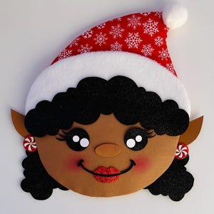 May include: A festive Christmas elf face decoration. The elf has black curly hair, brown skin, and a red and white Santa hat with snowflake patterns. The elf is wearing red and white candy earrings and has a red glittery smile.