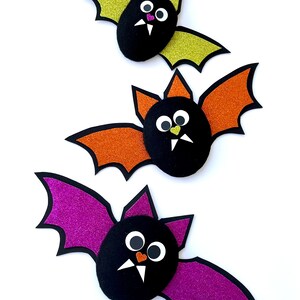 Könnte beinhalten: Drei Papierfledermäuse mit Glitzerflügeln. Die Fledermäuse sind schwarz mit weißen Augen und orangefarbenen oder rosafarbenen Nasen. Die Flügel sind grün, orange und lila.