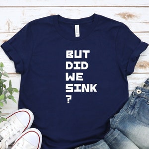 May include: A navy blue t-shirt with white text that reads "BUT DID WE SINK?" The shirt is laid flat on a white wooden surface with a pair of white sneakers and blue jeans.