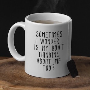 May include: White ceramic mug with the text "SOMETIMES I WONDER... IS MY BOAT THINKING ABOUT ME TOO?" in black. The mug is on a wooden surface, with steam rising from the contents. A wooden spoon is visible in the foreground.