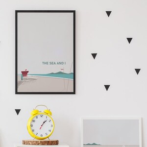 May include: A framed print featuring a minimalist illustration of a lifeguard stand on a beach with the text "THE SEA AND I". The print is hanging on a white wall with black triangle wall decals.
