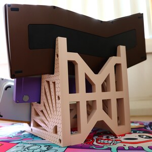 K&ouml;nnte beinhalten: Ein beigefarbener 3D-gedruckter St&auml;nder mit geometrischem Design, der eine braune und violette Tastatur h&auml;lt.