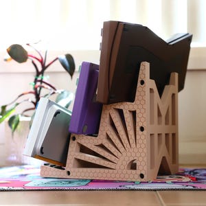 K&ouml;nnte beinhalten: Ein beigefarbener Plastikst&auml;nder mit einem Wabenmuster und einem Sonnenstrahl-Design h&auml;lt eine Sammlung von Videospielkassetten. Die Kassetten sind lila, grau und braun.