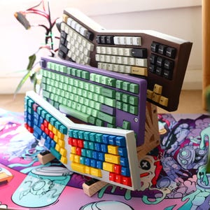 K&ouml;nnte beinhalten: Drei verschiedene Tastaturen, die auf einer bunten Schreibtischmatte gestapelt sind. Die oberste Tastatur ist braun mit wei&szlig;en und schwarzen Tasten. Die mittlere Tastatur ist lila mit gr&uuml;nen Tasten. Die unterste Tastatur ist wei&szlig; mit bunten Tasten.