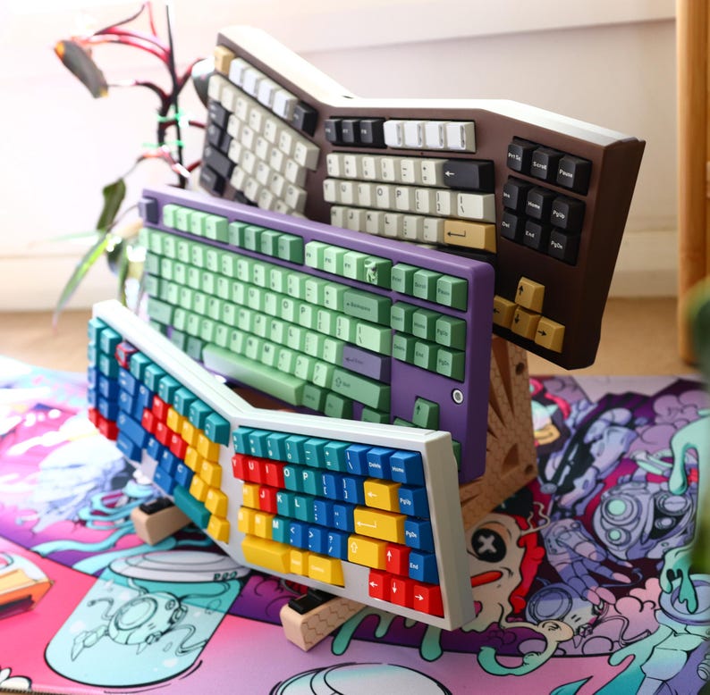 K&ouml;nnte beinhalten: Drei verschiedene Tastaturen, die auf einer bunten Schreibtischmatte gestapelt sind. Die oberste Tastatur ist braun mit wei&szlig;en und schwarzen Tasten. Die mittlere Tastatur ist lila mit gr&uuml;nen Tasten. Die unterste Tastatur ist wei&szlig; mit bunten Tasten.