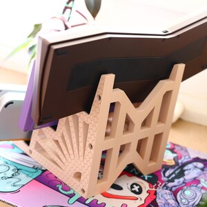 K&ouml;nnte beinhalten: Ein beigefarbener Plastikst&auml;nder mit geometrischem Design h&auml;lt eine braune Tastatur. Die Tastatur ist nach oben geneigt und der St&auml;nder steht auf einer bunten Oberfl&auml;che mit einem Cartoon-Design.