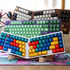 May include: Three different keyboards stacked on top of each other. The top keyboard has a green and purple colour scheme, the middle keyboard has a white and blue colour scheme, and the bottom keyboard has a white and brown colour scheme.