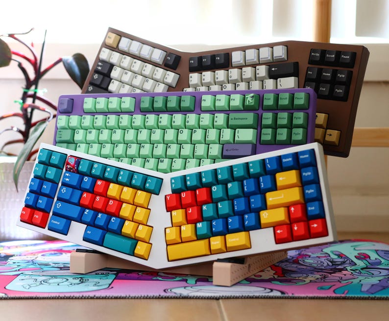 K&ouml;nnte beinhalten: Drei verschiedene Tastaturen, die &uuml;bereinander gestapelt sind. Die oberste Tastatur hat ein gr&uuml;n-violettes Farbschema, die mittlere Tastatur ein wei&szlig;-blaues Farbschema und die unterste Tastatur ein wei&szlig;-braunes Farbschema.