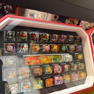 May include: A display case filled with colorful, square-shaped keycaps featuring various cartoon character designs. The keycaps are arranged in rows within a red and white framed case, with a clear cover.